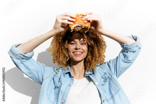 Obraz na płótnie Cheerful woman with curly hair holding a burger, showcasing a fun food concept in a bright, minimalist setting Emphasizing joy and indulgence in fast food w salonie