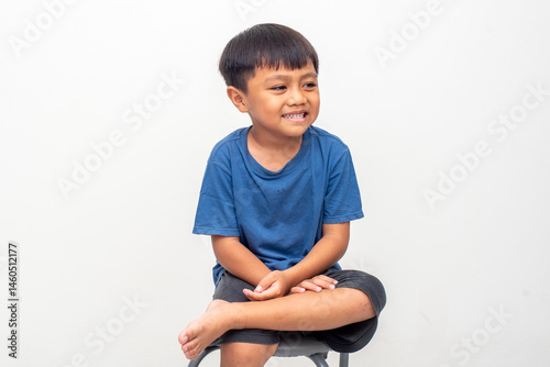A young boy sits cross-legged on a stool, wearing a blue shirt and shorts, smiling slightly against a plain white backdrop. Image toned and noise added, soft focus