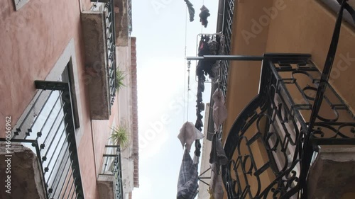 Colorful clothing items gently swinging between weathered buildings, capturing traditional domestic lifestyle on narrow wrought iron balcony in historic lisbon neighborhood