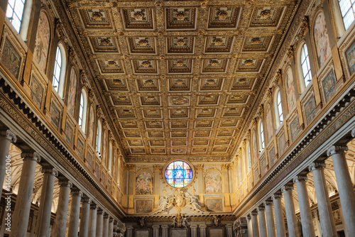 Ornate golden coffered ceiling and richly decorated interior of Basilica of Saint Mary Major in Rome Italy with rows of pews marble floor and illuminated altar at far end. High quality photo