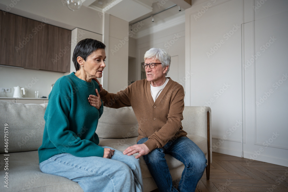 Obraz premium Shocked woman reacts to heartbreaking news, grabs chest in anxious heart attack while worried man holds close on home sofa. Emotional overload, sorrow and tragic news, breathing difficulty tight chest