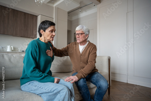 Shocked woman reacts to heartbreaking news, grabs chest in anxious heart attack while worried man holds close on home sofa. Emotional overload, sorrow and tragic news, breathing difficulty tight chest photo