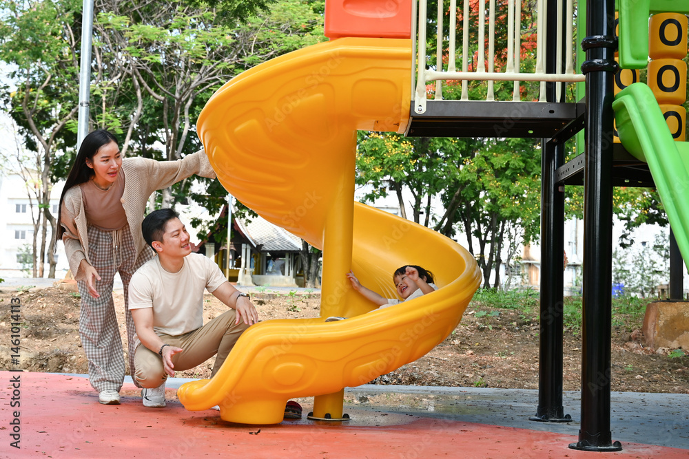 A family is standing in front of a slide. The child is sitting on the slide while the parents look on. Scene is happy and playful.