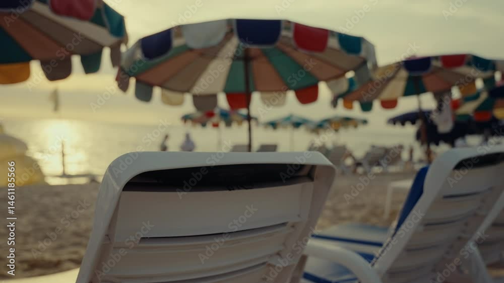 White beach chairs stand beneath vibrant, multi-colored umbrellas on a sandy beach in thailand, as the sun sets over the ocean, creating a tranquil and inviting scene in slow motion