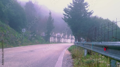 Static shot of wet road turn ending in a cemetery, Nature and woods around it. A bit of wind. Foggy weather.