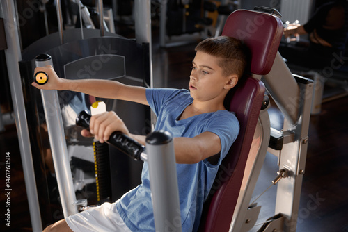 Teen boy doing sports exercise at machine for chest muscles pumping