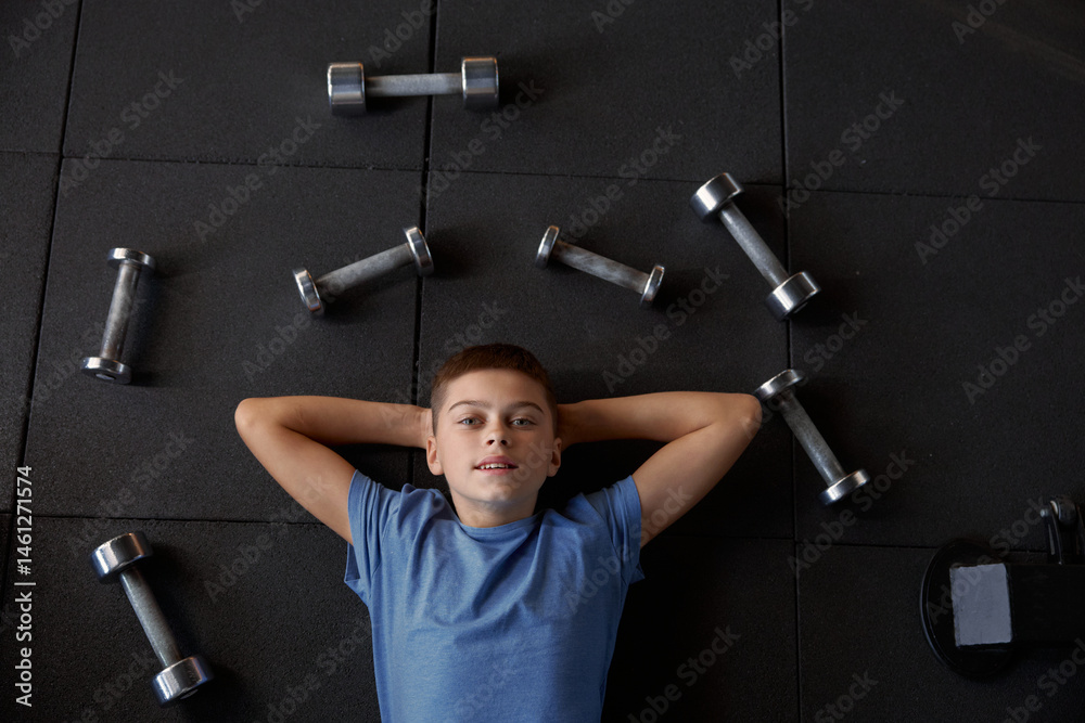 Overhead portrait view on boy lying on gym floor surrounded by dumbbells