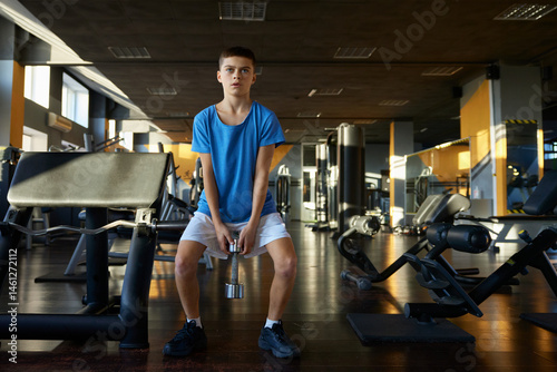 Athletic teenage boy squatting with dumbbells training over gym interior