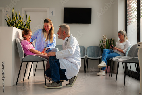 Senior male doctor engaging in conversation with a smiling girl and her mother, while another patient is using a phone in the bright, modern waiting room of a medical clinic