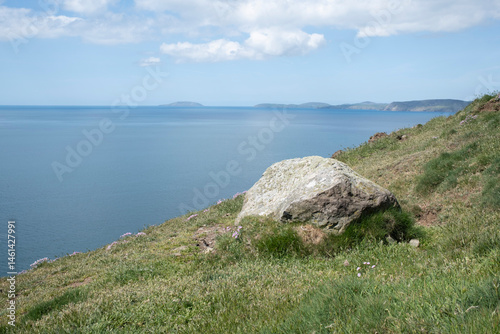 A boulder on green grass with flowers leads across a calm blue sea to a headland and Bardsey Island under a blue sky with cloud on the Lleyn Peninsular, Wales, UK.