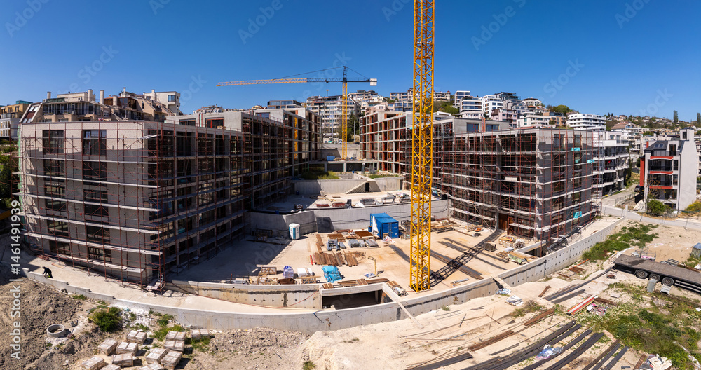 Fototapeta premium Panoramic view of a modern residential construction site with scaffolding, cranes, and unfinished buildings under a clear blue sky in an urban environment