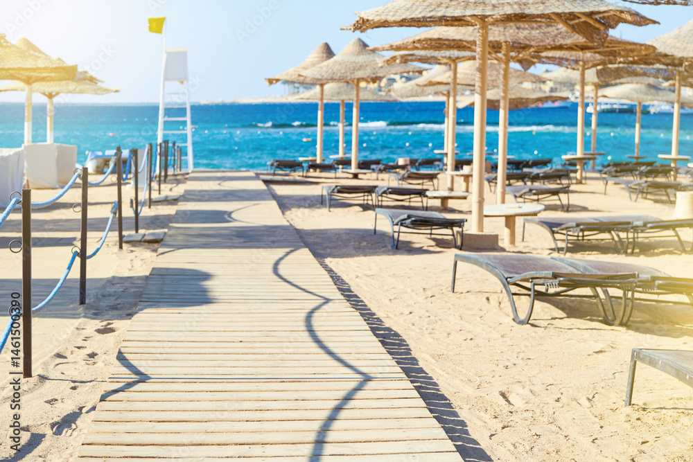 Fototapeta premium Beach umbrellas parasol made of wood and sunbeds standing on seashore against the backdrop of sunny blue sky and glare. View over tropical beach with rows of parasols. Vacation and Travel Concept.