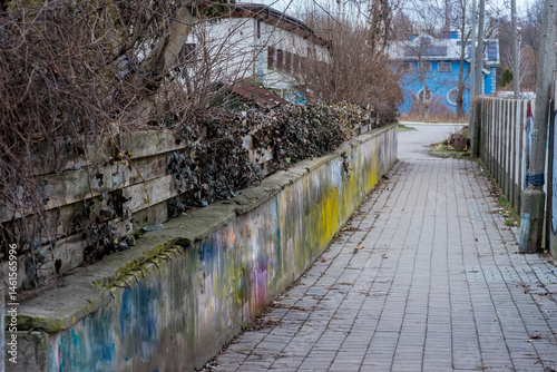 rochę zapomniana ulica w mieście wczesną wiosną. Wąska, brukowana ulica otoczona betonowymi murami, częściowo pokrytymi zwietrzałym graffiti. Rzadka roślinność, w tym nagie krzewy i pnący bluszcz.