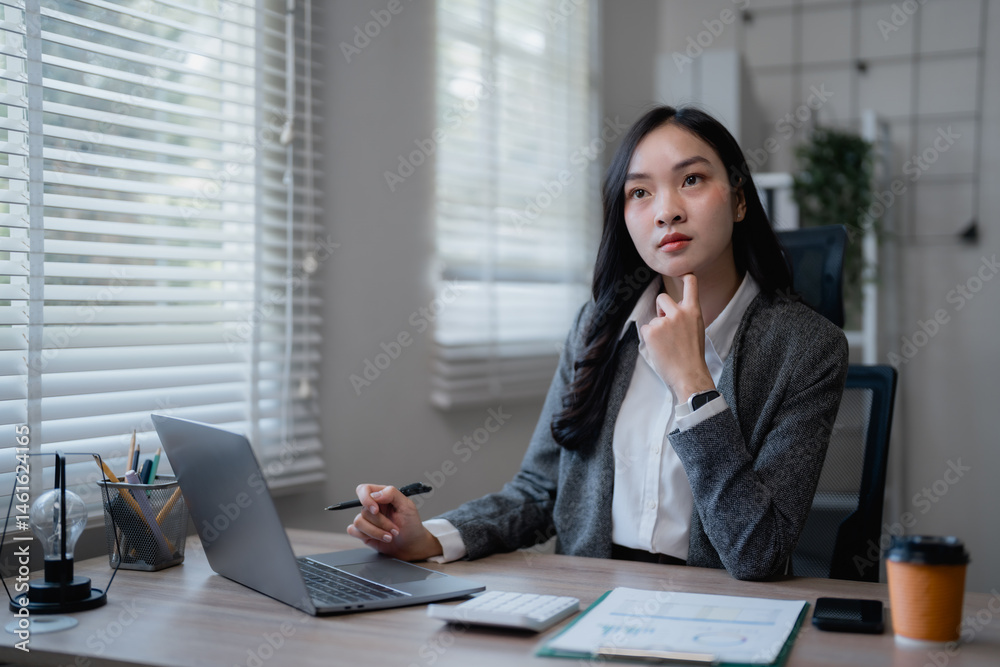 Young asian businesswoman is thinking and working using laptop computer and holding pen to analyze financial chart documents in modern office Young asian businesswoman is thinking and working using laptop computer and holding pen to analyze financial chart documents in modern office