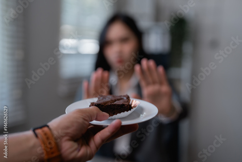 Businesswoman is raising her hands and refusing a chocolate brownie offered by a colleague, showing her commitment to a healthy lifestyle or dietary restrictions