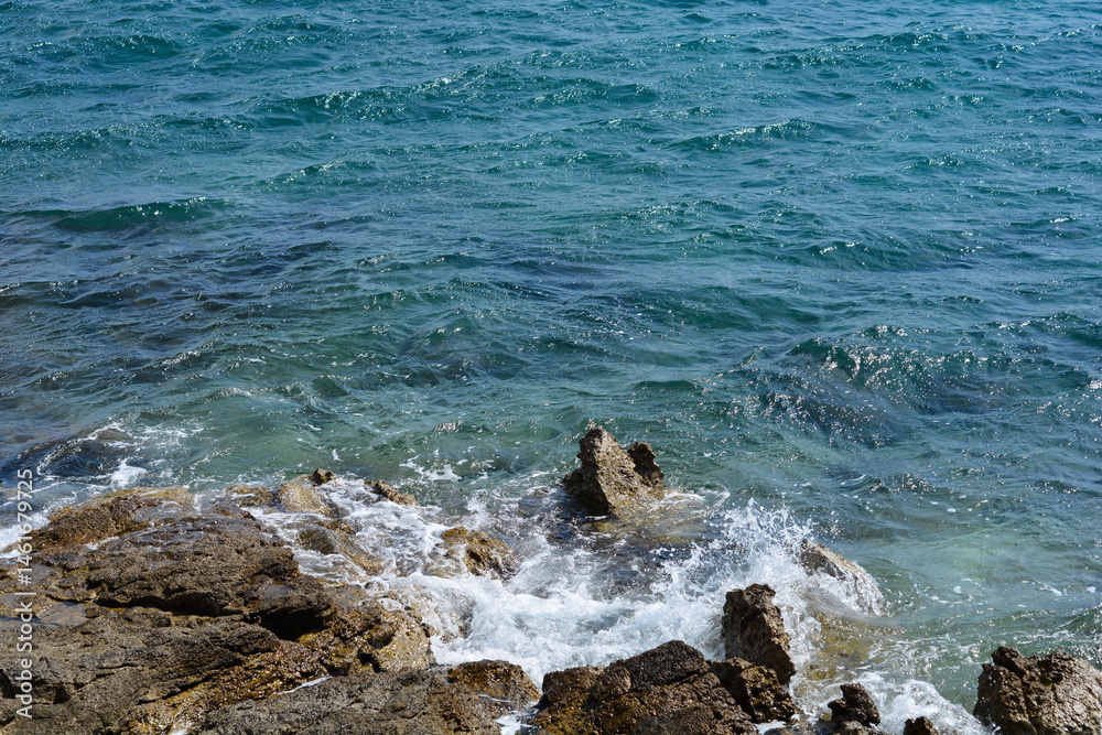 Dark blue sea waves crash against rugged, dark rocks along the shore, creating white foamy splashes under natural light