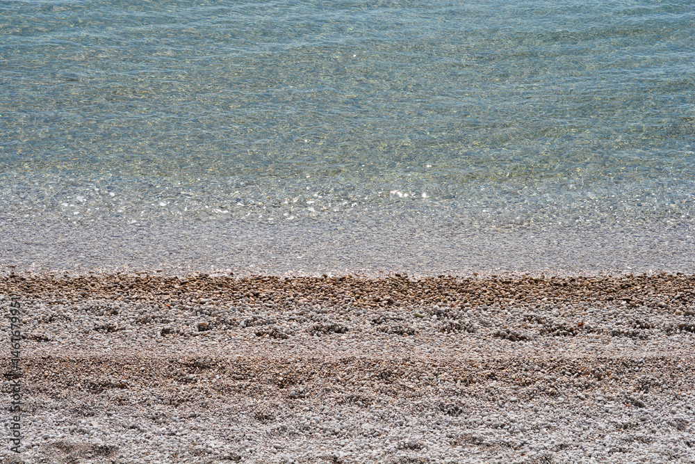 Clear turquoise sea gently washes over a pebble beach under a bright sky