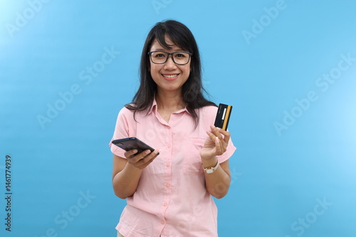Asian woman happy using mobile smart phone online internet financial banking payment purchasing e-commerce product filling a billing with credit debit card casual shirt isolated blue background