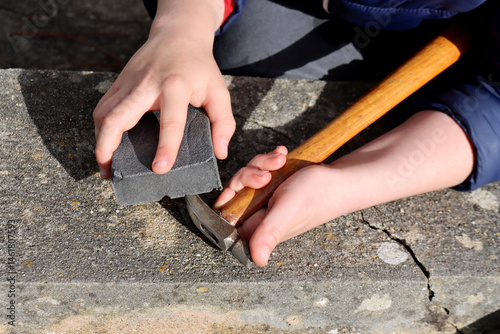 Child holding a hammer and sanding block, working on a stone surface outdoors. Concept of manual work, skill development, and hands-on learning.