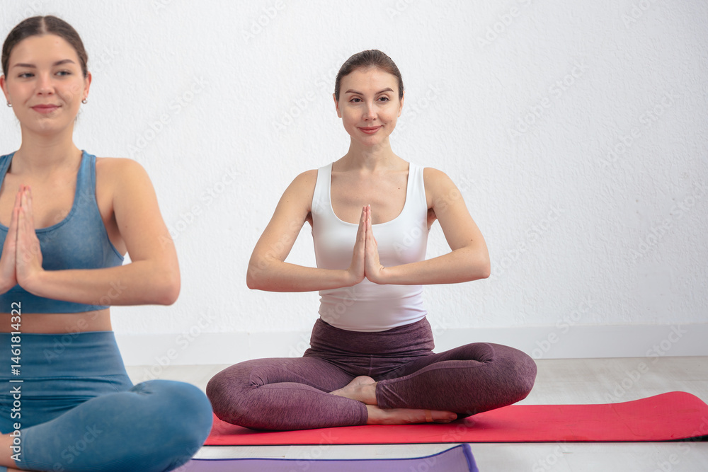 Fototapeta premium Smiling Caucasian women sitting in lotus pose with hands in prayer position,meditating on yoga mats indoors. Ideal for wellness, mindfulness, meditation, peace, and healthy lifestyle themes. 