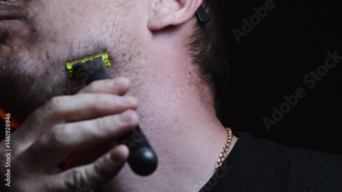 young man shaving his face with a trimmer for shaving his mustache and beard in a dark room 