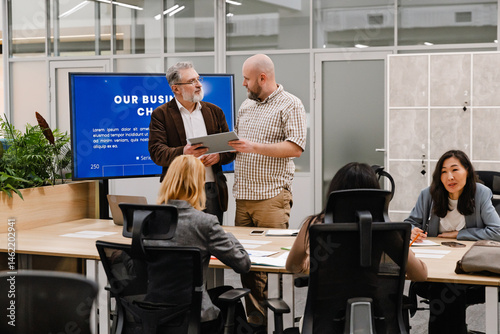 A diverse group of adult coworkers, including an elderly White man and an Asian woman, collaborate during a business meeting in a modern office. Two men stand near a large screen.