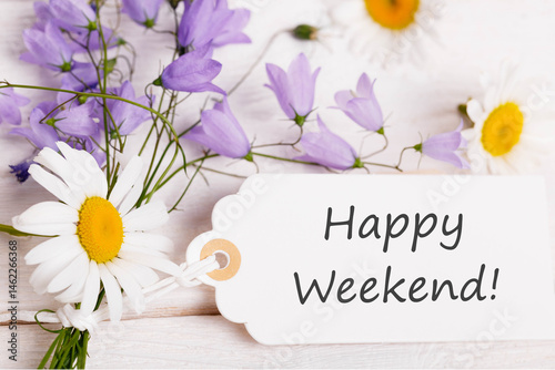 Happy weekend message on white gift tag with daisies and violet bellflowers on rustic wooden background, floral greeting card design, top view composition for summer celebration or weekend wishes