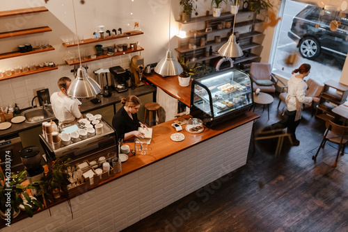 Papier peint A group of white adults, including baristas and waitstaff, prepare coffee and serve tables at a cozy cafe with wooden floors and a pastry display counter