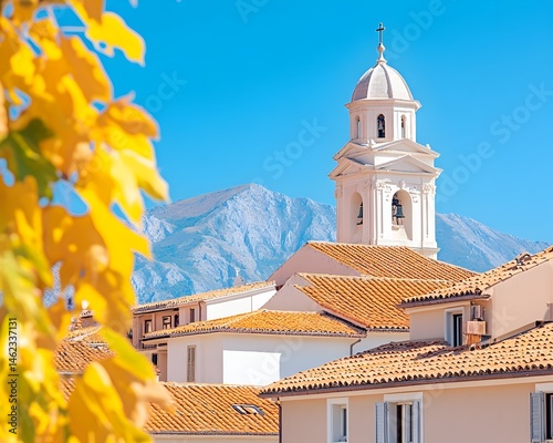 Obraz na płótnie Sunlit church steeple and terracotta rooftops nestled against a mountain backdrop, partially obscured by autumn leaves w salonie