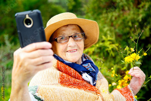 Portrait of a grandmother in the garden taking a photo of herself on her phone. An old woman takes a selfie on her phone against the backdrop of flowering bushes. The concept of old age, boarding hous