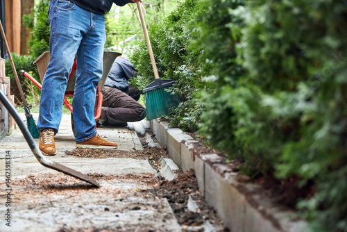 A gardener working in a summer garden, sweeping a path with a broom and cleaning up soil from plants
