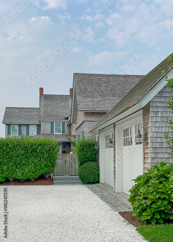 A beautifully maintained coastal home with cedar shingles, a gravel driveway, white double garage doors, lush greenery, and a bright blue sky dotted with soft clouds.