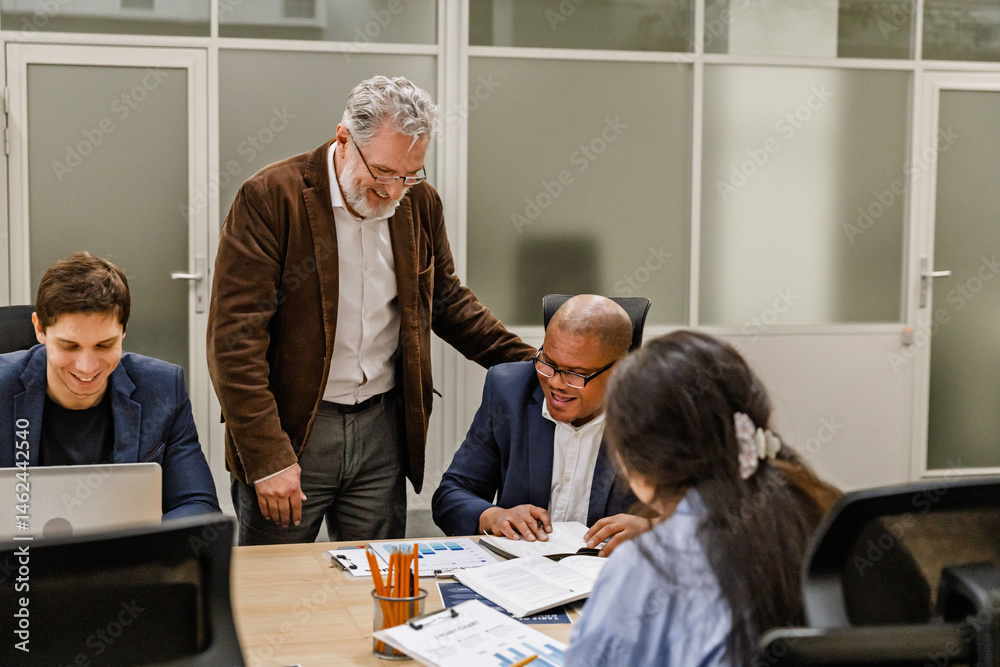 Obraz premium A male worker stands and looks at a clipboard on a table with a group of three workers sitting behind them, they are smiling, in a meeting
