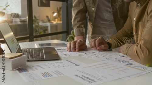 Midsection shot of senior manager sitting at white desk discussing financial documents with unknown male coworker standing next to him
