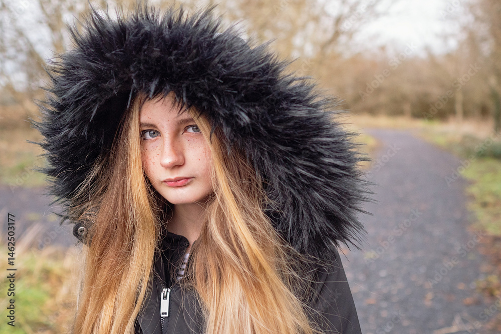 A girl with long blonde hair and a black hooded jacket in a park. She is looking at the camera. Selective focus. Fall or autumn cold season. Keep yourself warm and look stylish concept.