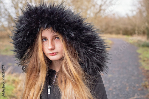 Obraz na płótnie A girl with long blonde hair and a black hooded jacket in a park. She is looking at the camera. Selective focus. Fall or autumn cold season. Keep yourself warm and look stylish concept. w salonie