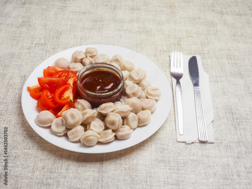 Russian dumplings on a white plate with brown sauce in a glass small jar and fresh cut tomatoes. Popular East European meal. Simple tasty food.