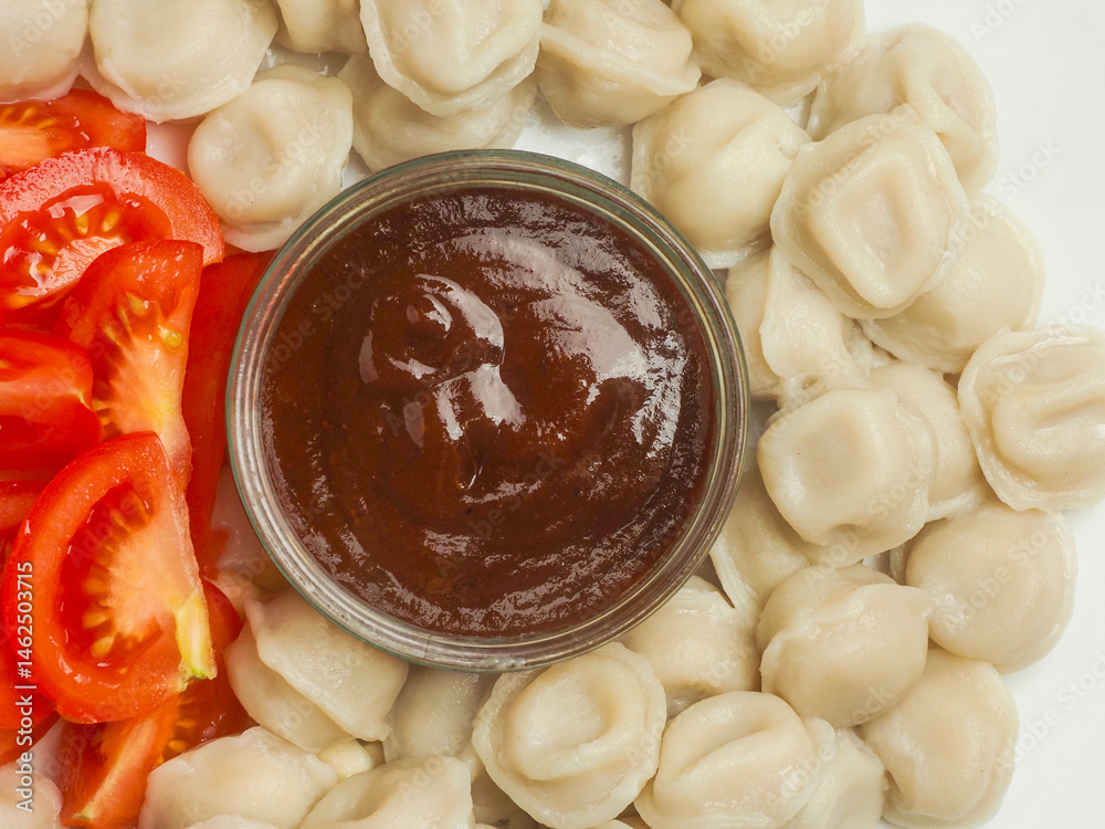 Russian dumplings on a white plate with brown sauce in a glass small jar and fresh cut tomatoes. Popular East European meal. Simple tasty food. Close up.