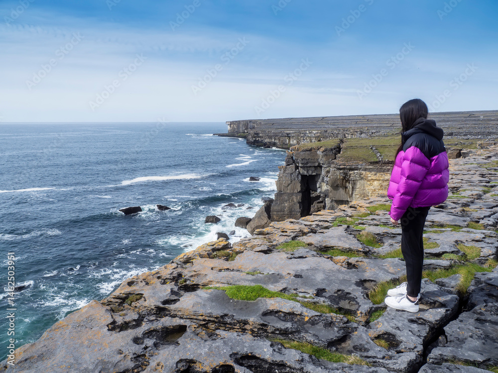 Teenager girl in purple jacket enjoys stunning nature of Aran island, county Galway, Ireland. Model is standing on edge of a cliff with view on the ocean and cloudy sky. Travel and tourism.