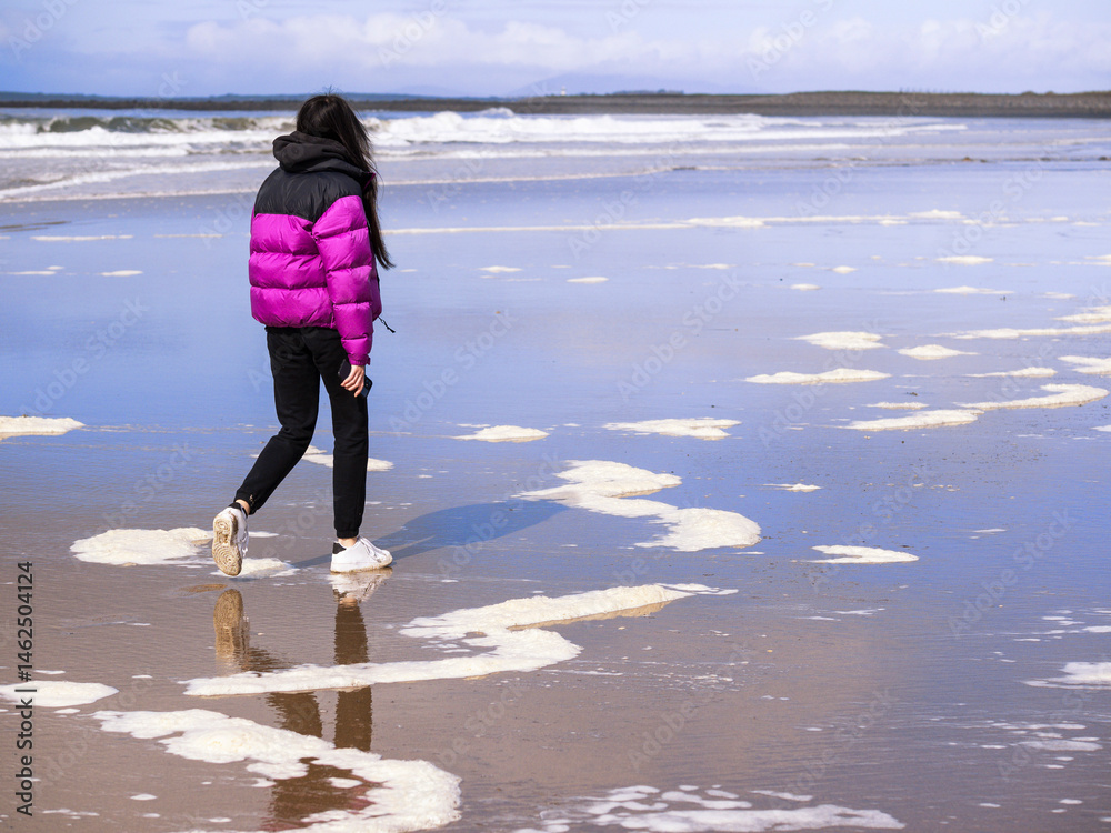 Teenager girl in purple jacket walking on a wet sandy beach which reflects blue cloudy sky. Travel to seaside concept. Relaxing activity by the ocean. Selective focus.