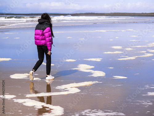 Obraz na płótnie Teenager girl in purple jacket walking on a wet sandy beach which reflects blue cloudy sky. Travel to seaside concept. Relaxing activity by the ocean. Selective focus. w salonie