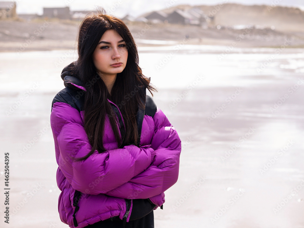 Portrait of a young teenager girl on a ocean beach with town in the background. Light and airy look. Model with long dark hair in purple jacket hands crossed, and sad face expression.