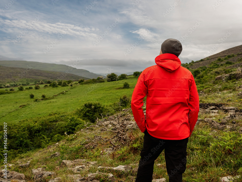 Man in red jacket looking at stunning nature scenery with mountains and dramatic sky. Burren, Ireland. Irish nature scene. Travel and tourism concept. Outdoor activity and sightseeing.