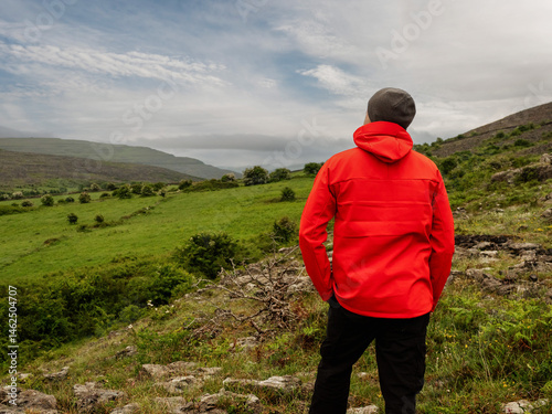 Obraz na płótnie Man in red jacket looking at stunning nature scenery with mountains and dramatic sky. Burren, Ireland. Irish nature scene. Travel and tourism concept. Outdoor activity and sightseeing. w salonie