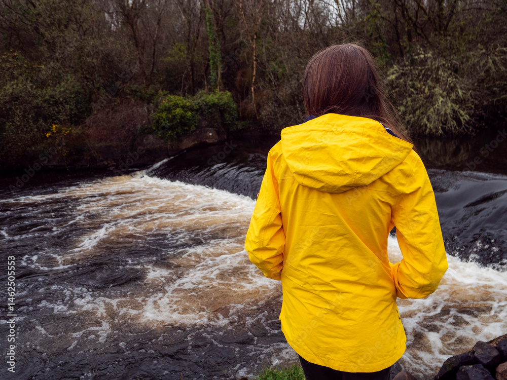 Female tourist in yellow jacket looking at a small river with waterfall. Travel and tourism. Enjoy nature scenery theme. Girl standing by a river with nice view.