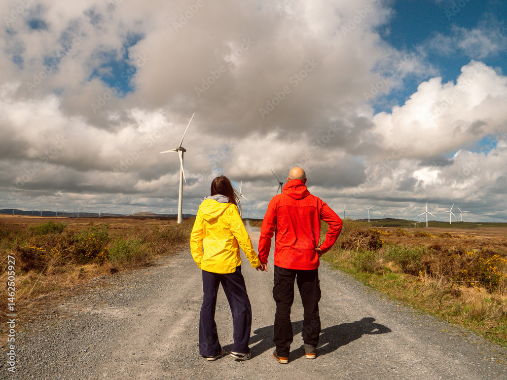 Father with daughter on a road and field with wind turbines and vast field. Warm sunny day with blue cloudy sky. Man in red jacket, girl in yellow. Explore nature and technology concept.
