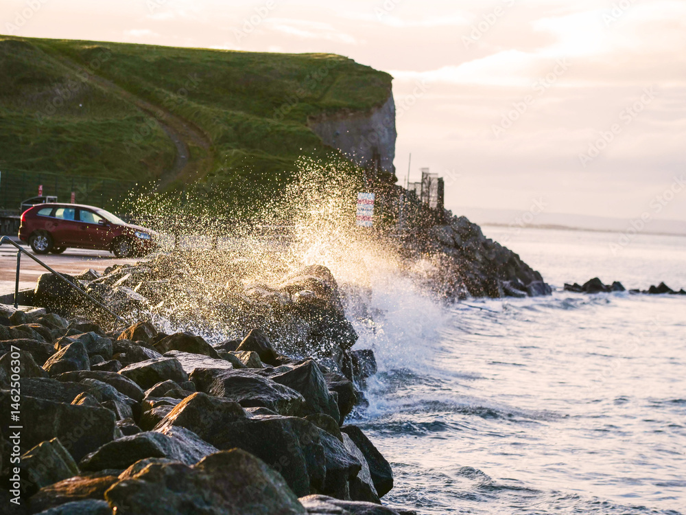 Wave crushes on big rocks next to a car park with cars, cliff in the background. Calm and peaceful sunset light. Silverstrand area, Galway city, Ireland. Popular tourist area with stunning scenery.