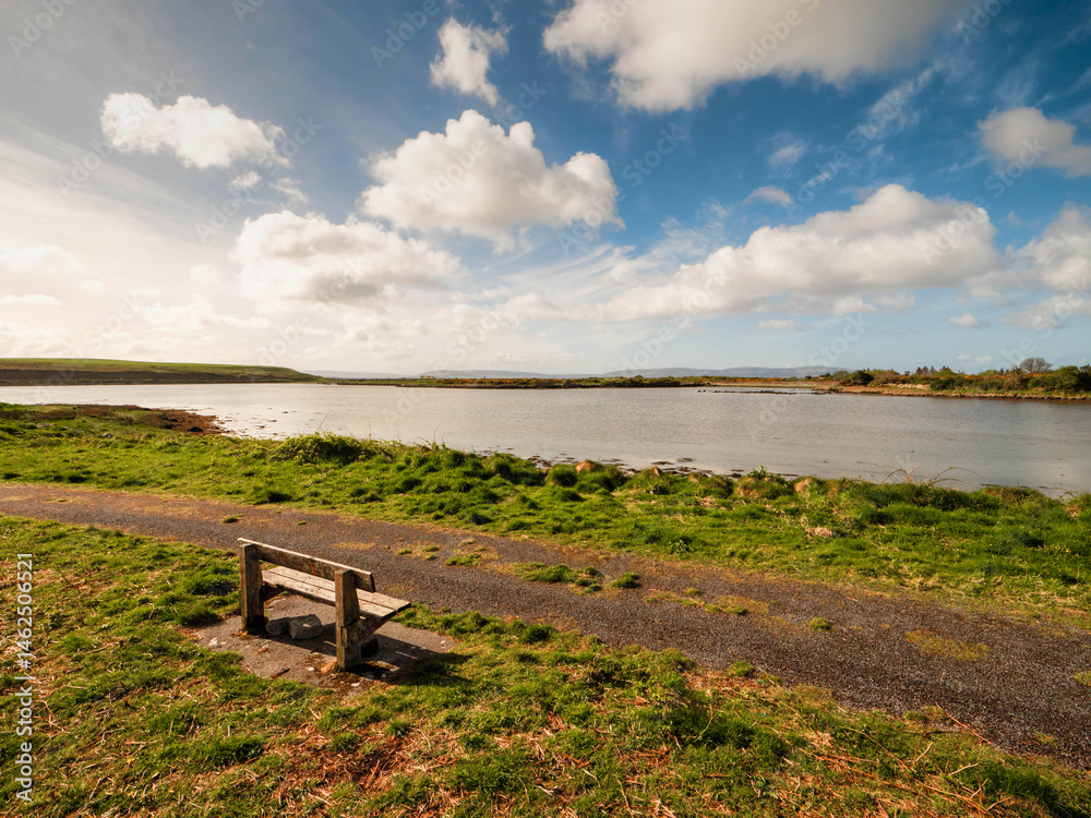 One wooden old bench by a walking path with stunning view on open space with lake and amazing blue cloudy sky. Nature scenery in a park and great place to relax and enjoy the view.