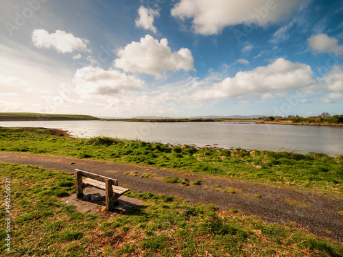 Obraz na płótnie One wooden old bench by a walking path with stunning view on open space with lake and amazing blue cloudy sky. Nature scenery in a park and great place to relax and enjoy the view. w salonie