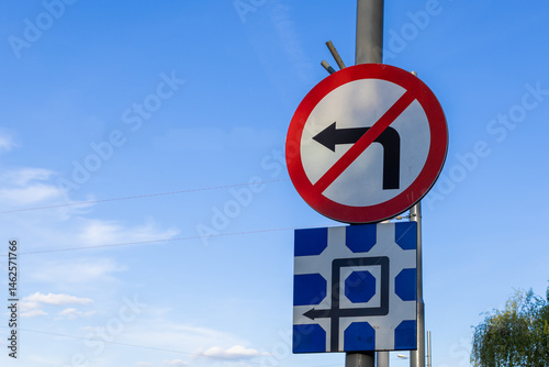 Two traffic signs are mounted on a pole against blue sky, including a No Left Turn symbol and complex diagrammatic sign for road regulation driver guidance